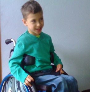 Zdjęcie przedstawia chłopca siedzącego na wózku w zielonym sweterku