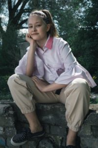 Zdjęcie przedstawia dziewczynę siedzącą w różowej koszuli i beżowych spodniach