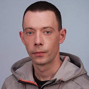 Zdjęcie przedstawia portret mężczyzny o krótkich, ciemnobrązowych włosach i jasnych oczach. Mężczyzna patrzy bezpośrednio w stronę aparatu, ma neutralny wyraz twarzy. Ubrany jest w szarą bluzę z kapturem i czerwonymi detalami oraz czarną koszulkę pod spodem. W tle znajduje się gładka, jasnoszara powierzchnia. Zdjęcie wykonano w ujęciu portretowym, obejmującym głowę i górną część tułowia, przy równomiernym oświetleniu.