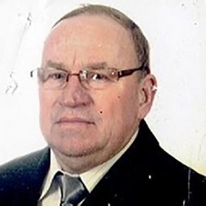 Zdjęcie przedstawia starszego mężczyznę o krótkich, siwiejących włosach, noszącego prostokątne okulary w brązowych oprawkach. Mężczyzna ma na sobie ciemny garnitur, jasną koszulę i ciemny krawat. Patrzy lekko w bok z poważnym, spokojnym wyrazem twarzy. W tle znajduje się jasna ściana z delikatnymi śladami i nierównościami. Portret wykonano z bliska, obejmując głowę, ramiona i górną część tułowia mężczyzny.