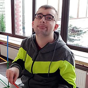 Mężczyzna w czarnych okularach i szarej bluzie z kapturem, ozdobionej jaskrawożółtymi i czarnymi pasami, siedzi na wózku. Ma krótkie, ciemne włosy i lekki zarost. Patrzy w stronę aparatu z delikatnym uśmiechem. W tle widać duże okna, kaloryfer oraz fragment pomieszczenia z wyposażeniem sportowym.