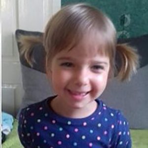 Zdjęcie przedstawia uśmiechniętą dziewczynkę z jasnobrązowymi włosami, związanymi w dwa kucyki po bokach głowy, oraz prostą grzywką. Dziecko ma na sobie granatową bluzkę z kolorowymi kropkami. Patrzy w stronę aparatu i szeroko się uśmiecha. Siedzi na kanapie z szarymi poduszkami. W tle widoczna jest ciemnozielona powierzchnia oraz fragment jasnej ściany. Zdjęcie przedstawia dziewczynkę w domowym otoczeniu, w kadrze obejmującym głowę i górną część tułowia.